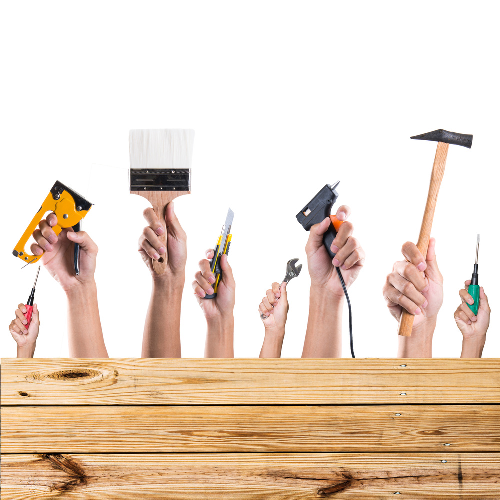 A group of hands holding various tools on top of a wooden board preparing to DIY.